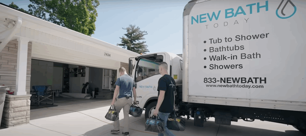 Two bathroom remodel technicians walking toward a home carrying tools beside a New Bath Today service truck